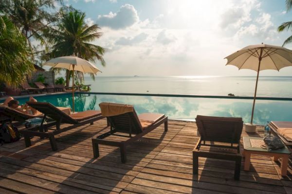 A tropical poolside scene with lounge chairs, umbrellas, and palm trees overlooks a calm body of water under a bright sky.