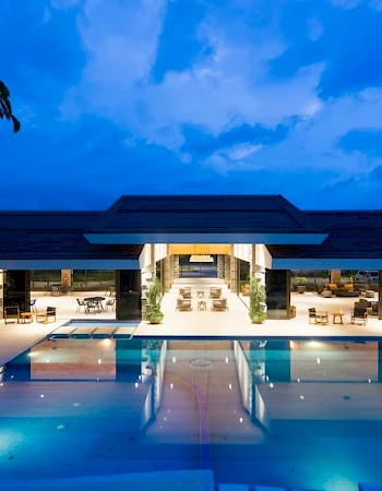 The image shows a modern building at dusk with a lit-up swimming pool in the foreground, reflecting the evening sky and structure.