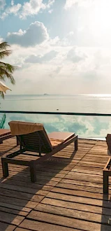 Poolside loungers and umbrellas overlook a serene ocean view under a partly cloudy sky, with palm trees enhancing the tropical setting.