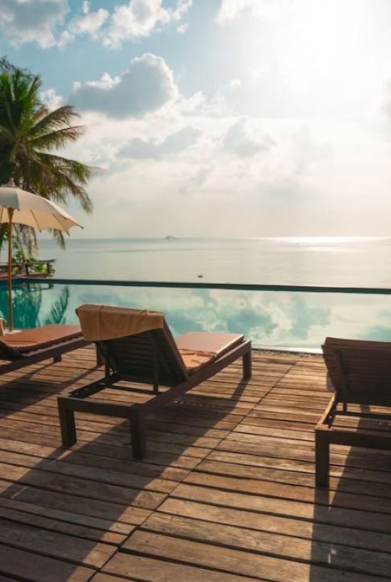 Chairs and umbrellas by a pool, with a view of the ocean and palm trees under a bright sky.