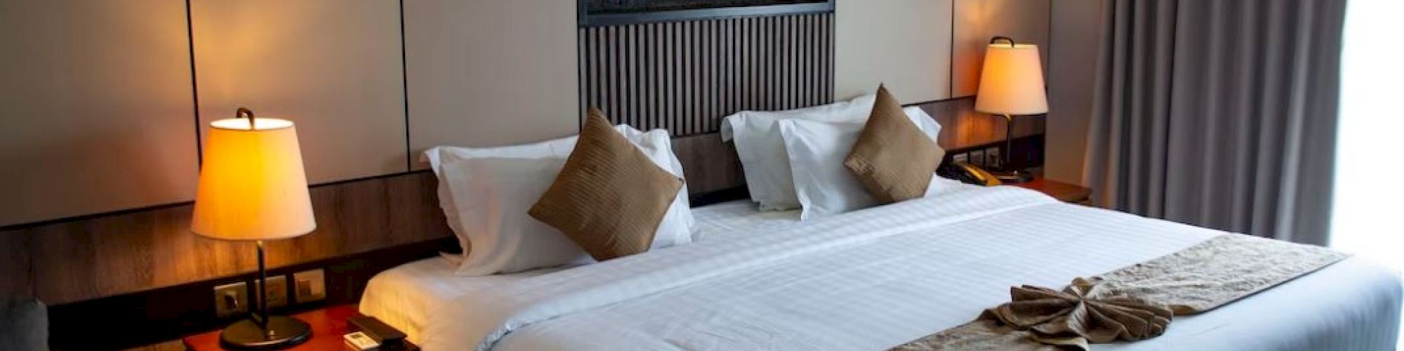 A modern hotel room with a large bed, decorative pillows, lamps, and a wall-mounted fireplace.