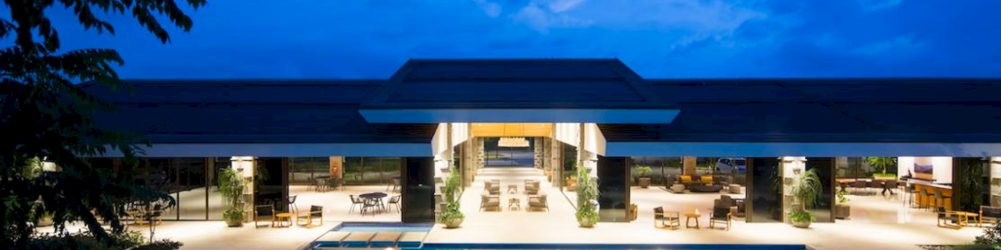 A modern building with a large swimming pool in front, beautifully lit at night, under a cloudy blue sky.