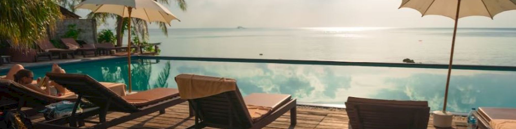 A tranquil poolside scene with lounge chairs, umbrellas, palm trees, and ocean views under a bright sun and cloudy sky.