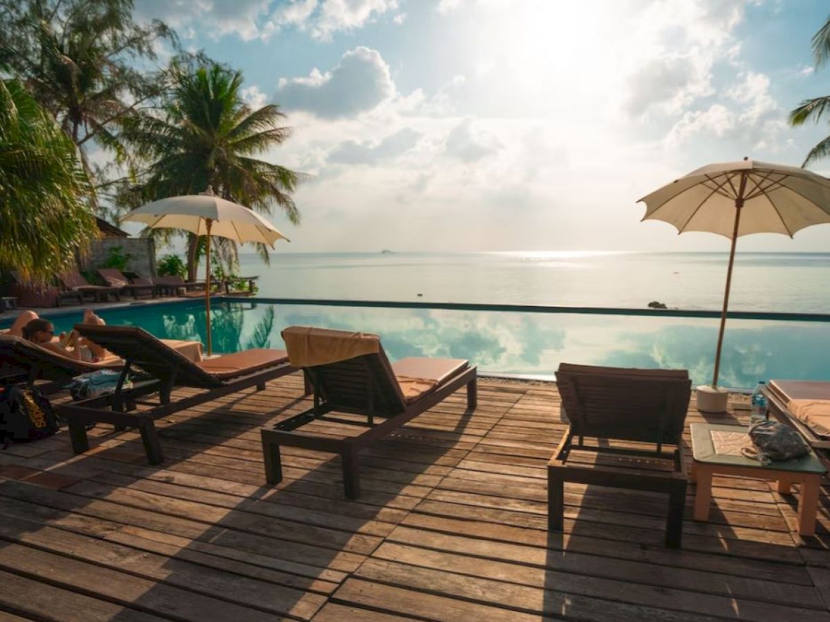 A tranquil poolside scene with lounge chairs, umbrellas, palm trees, and a serene ocean view under a partly cloudy sky.