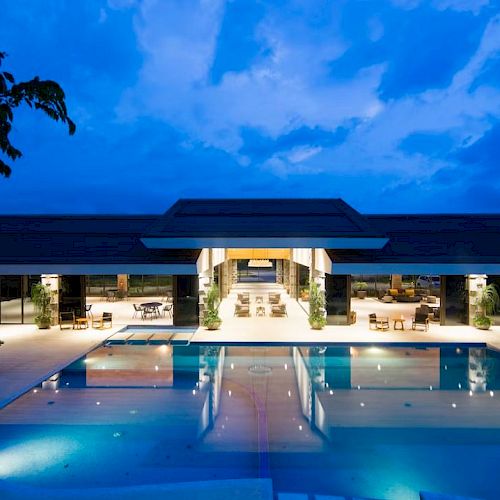 A luxurious modern building with an illuminated swimming pool at dusk, featuring open architecture and a serene ambiance.