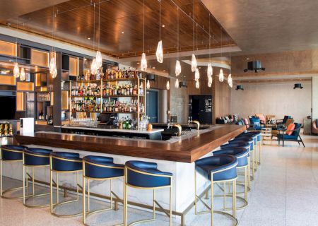 The image shows a modern bar with a central counter, blue stools, and a well-stocked back shelf. Pendant lights hang from the ceiling.