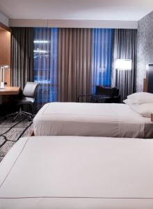 The image shows a modern hotel room with two neatly made beds, a desk, chair, TV, and stylish lighting and decor.