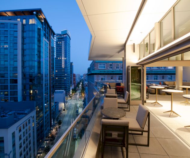 A modern cityscape at dusk features a rooftop terrace with seating, overlooking illuminated buildings and a street below.