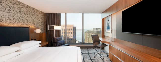 The image shows a modern hotel room with a large bed, wall-mounted TV, desk, and a window offering a city view.