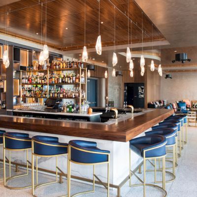 A modern bar interior with a central island, stools, hanging lights, and a well-stocked shelf of bottles in a stylish, open space.