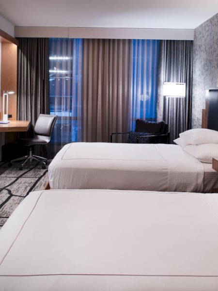 The image shows a hotel room with two beds, a large TV, a desk and chair, modern lamps, and patterned curtains and wallpaper.