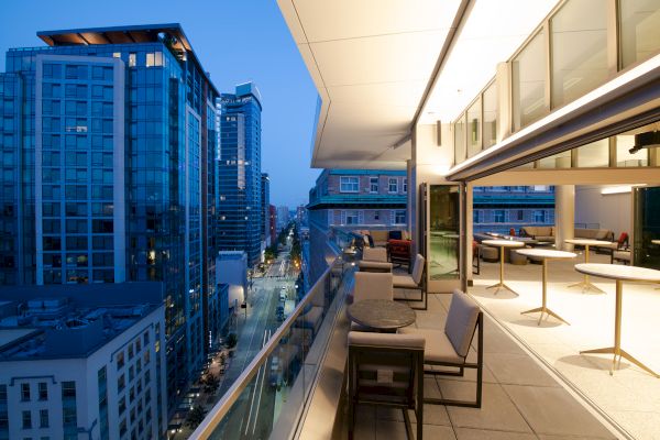 A city view from a modern balcony at dusk, showcasing tall buildings and street lights with seating and tables on the terrace.