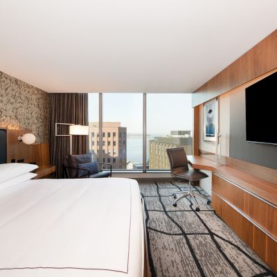 Modern hotel room with a large bed, wall-mounted TV, desk chair, and a window view of city buildings and water.