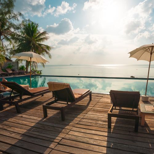 A serene poolside scene with lounge chairs, umbrellas, and palm trees overlooking a tranquil ocean under a sunny sky.