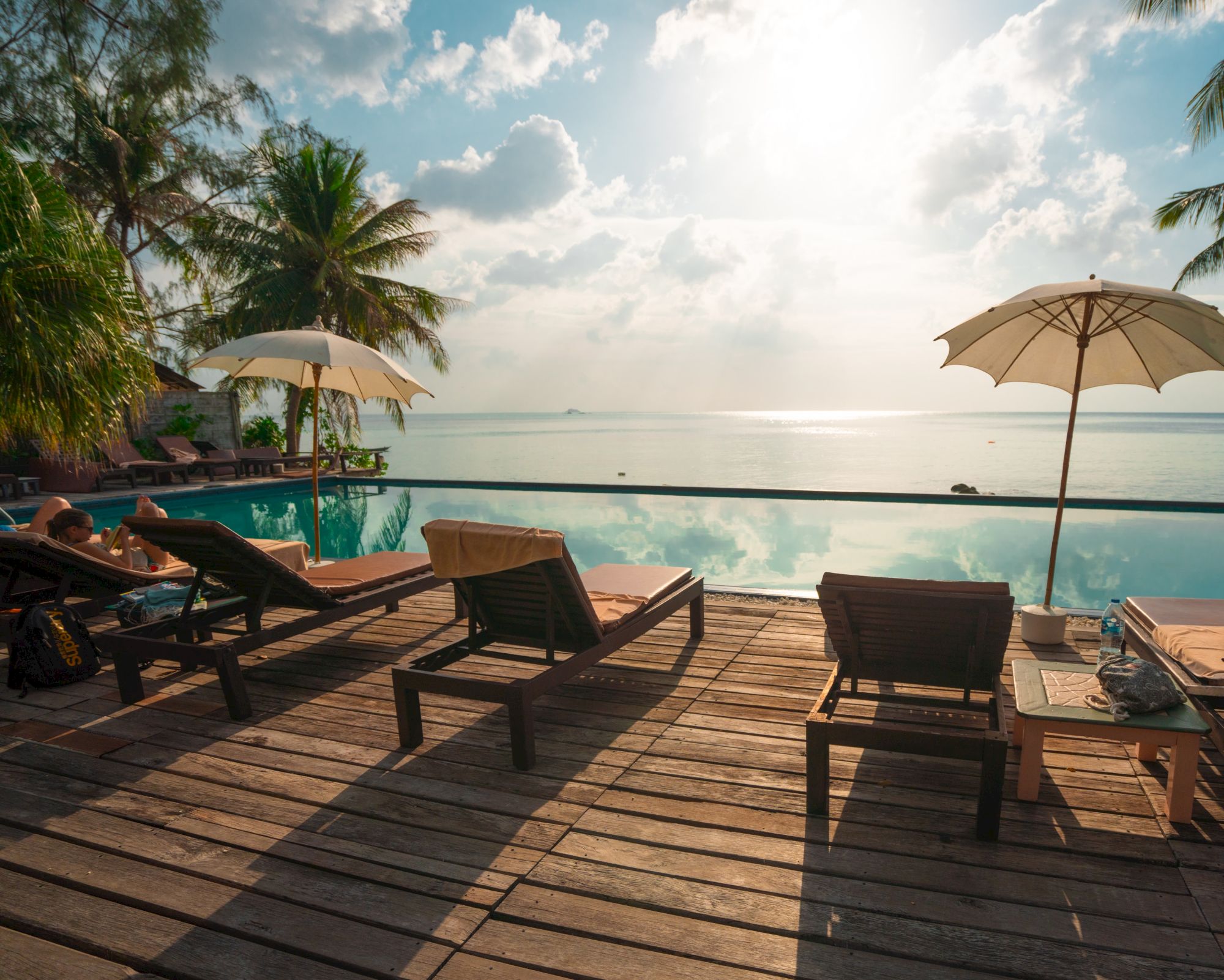 A serene ocean view with a wooden deck, lounge chairs, umbrellas, and a swimming pool, under a partly cloudy sky and bright sun.