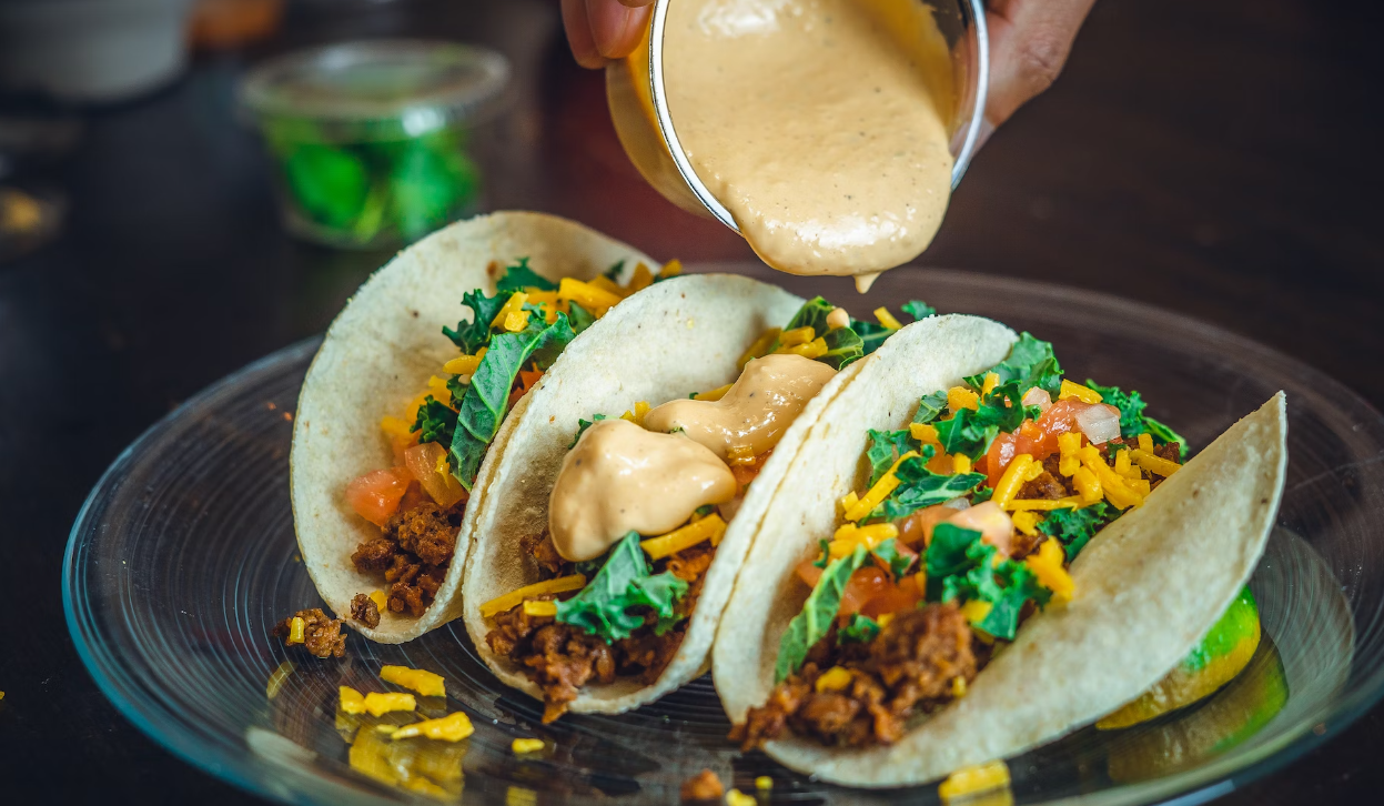 Three tacos filled with meat, cheese, tomatoes, and greens on a plate, with a sauce being poured over them from a cup.