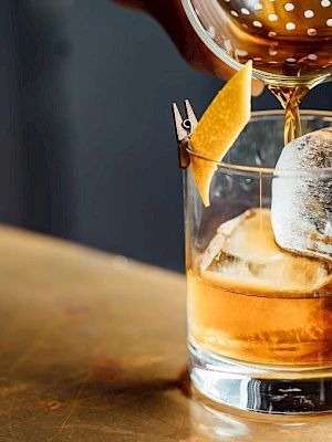 A cocktail is being poured into a glass with a large ice cube and a twist of citrus peel garnish.