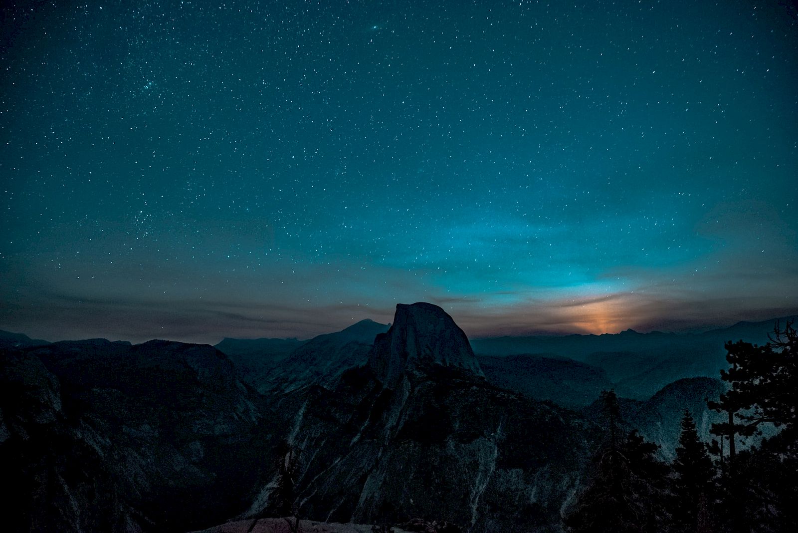 A starry night sky over a mountainous landscape with a glowing horizon, featuring a prominent, dark silhouette of a peak.