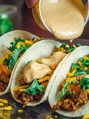 Three tacos filled with meat, cheese, lettuce, and tomatoes are being topped with a creamy sauce.