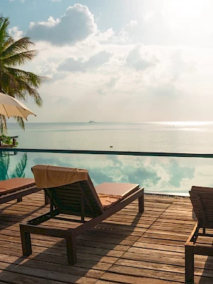 A serene tropical setting with lounge chairs and umbrellas by an infinity pool, overlooking a calm ocean under a partly cloudy sky.