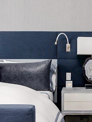 A modern bedroom with a navy and white color scheme, featuring a bed, nightstand, lamp, and wall-mounted reading light.