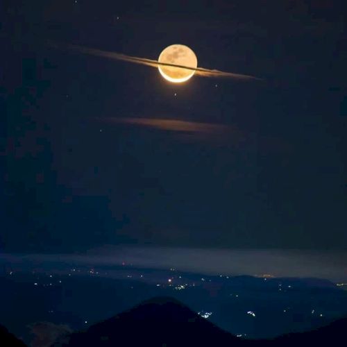 A full moon appears with clouds that look like Saturn's rings, above a landscape with scattered lights in the distance.