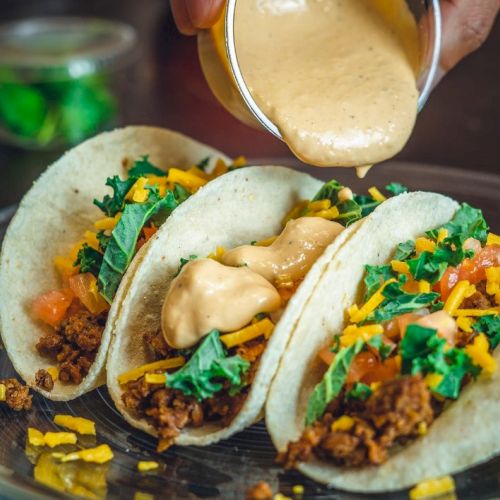 Tacos filled with meat, shredded cheese, lettuce, tomato, and a sauce being poured on top, served on a clear plate.