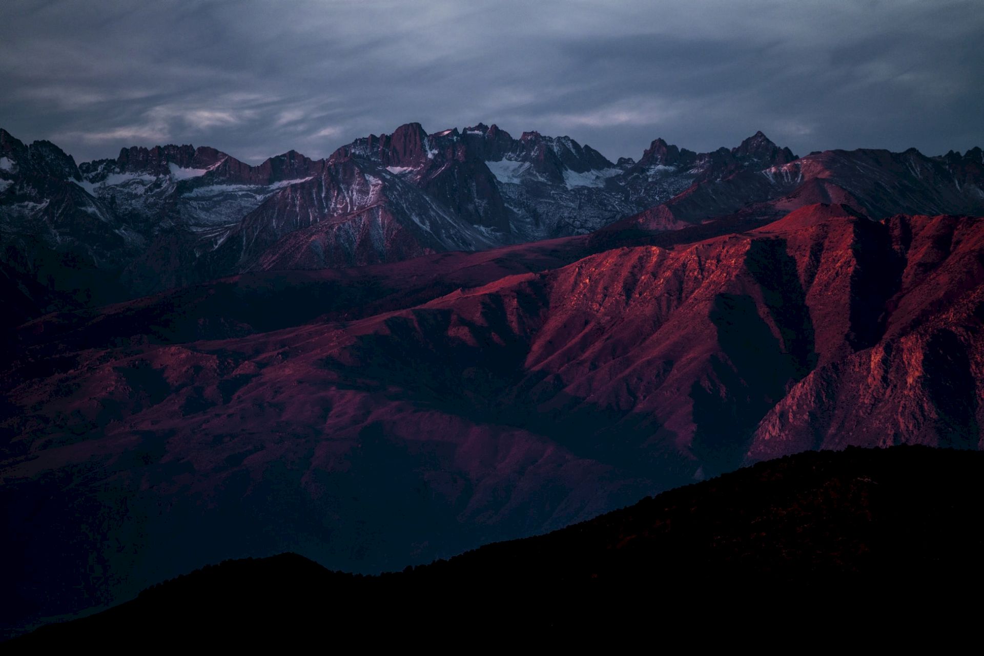 A mountain range is bathed in red sunlight, with dramatic, cloudy skies enhancing the scene's contrast and depth.