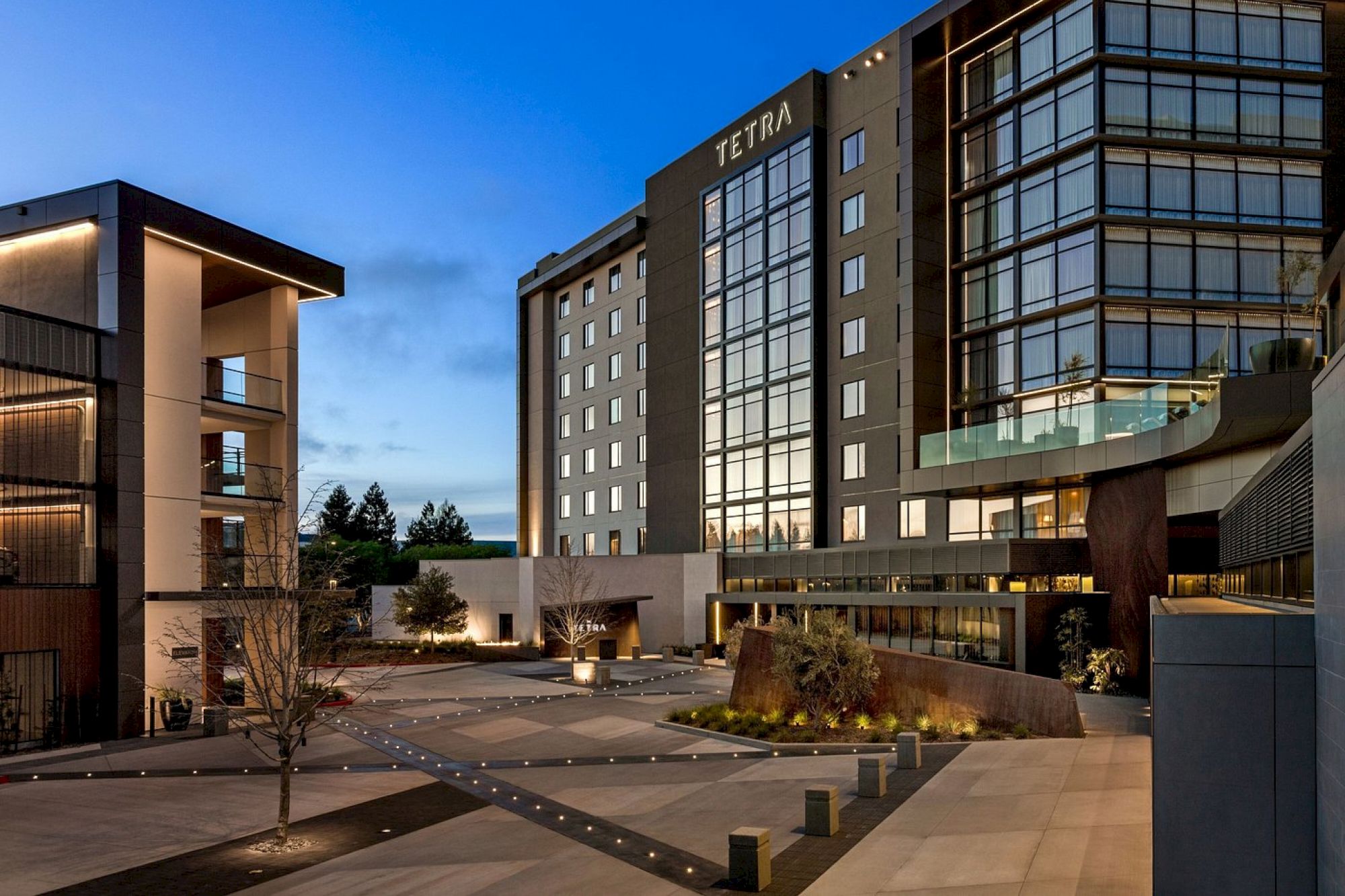 The image shows a modern hotel complex at dusk, featuring sleek architecture, large windows, and a well-lit courtyard area.