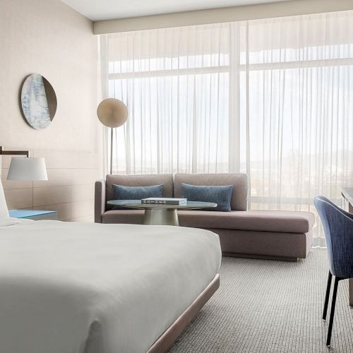 A modern hotel room with a bed, sofa, desk, and large window with sheer curtains, creating a bright and inviting atmosphere.