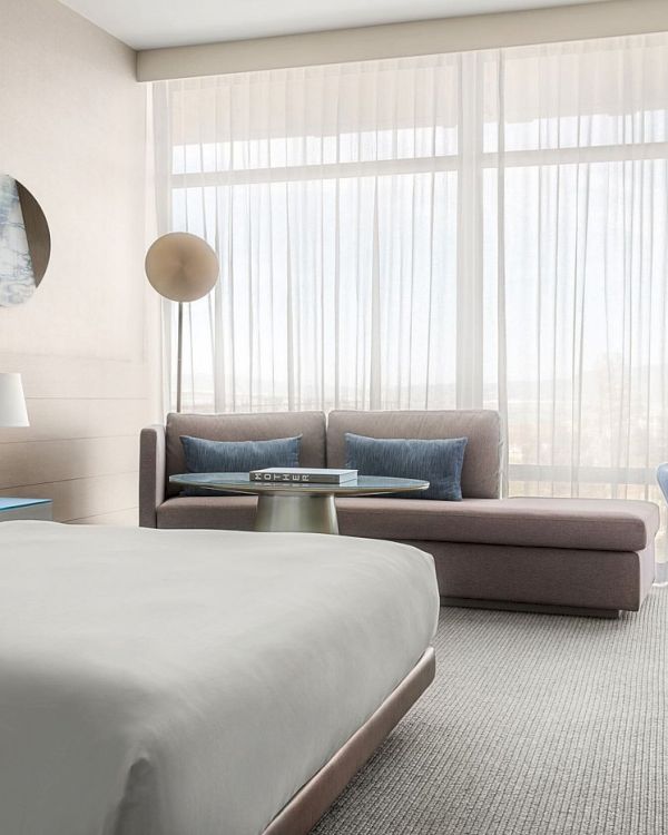 A modern hotel room with a bed, sofa, desk, and large window with sheer curtains, creating a bright and inviting atmosphere.
