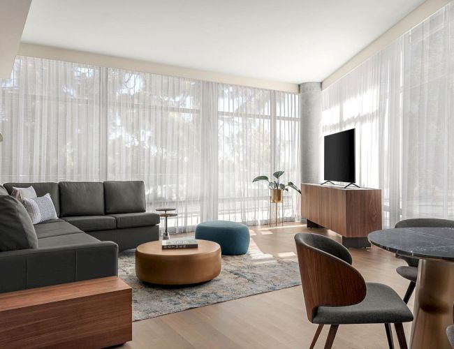 A modern living room with a sectional sofa, TV on a stand, round coffee table, and dining set, surrounded by large windows with sheer curtains.