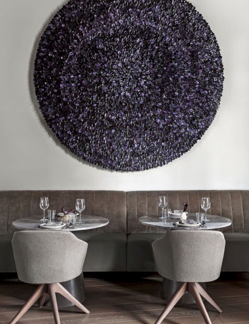 Two small round tables with chairs in a modern setting, and a large circular textured wall art piece above them.