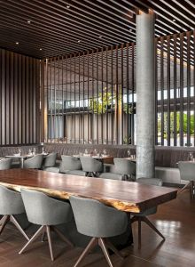 The image shows a modern, stylish restaurant interior with wooden accents, elegant gray chairs, and a large communal table.