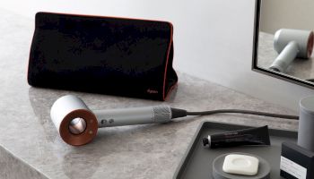 The image shows a hairdryer on a countertop next to a black travel bag, a tray with toiletries, and a partial view of a mirror.