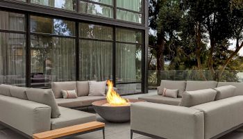 Outdoor seating with gray sofas and a fire pit on a patio near a modern building with large windows surrounded by trees.