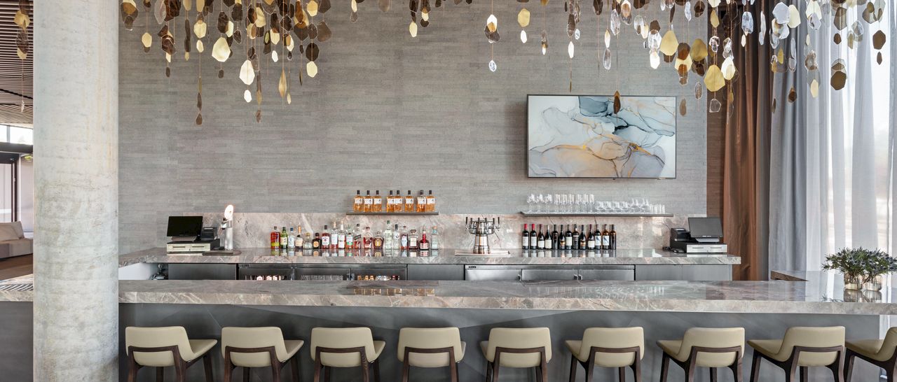 A modern bar with many bottles, backlit counters, tall stools, and glowing gold hanging pendant lights overhead.
