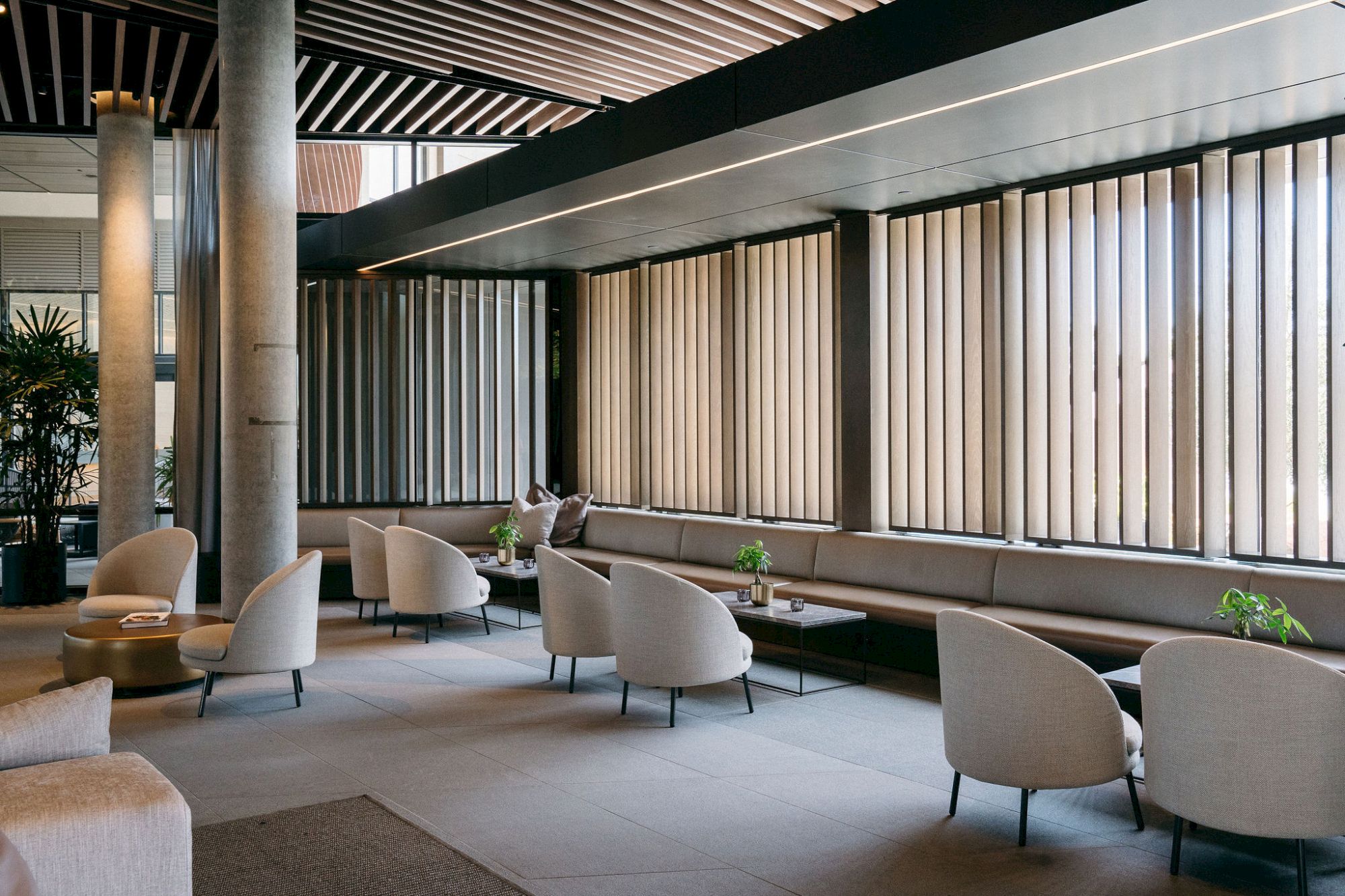 The image shows a modern lounge with comfortable chairs, tables, and large vertical blinds, creating a relaxed atmosphere.