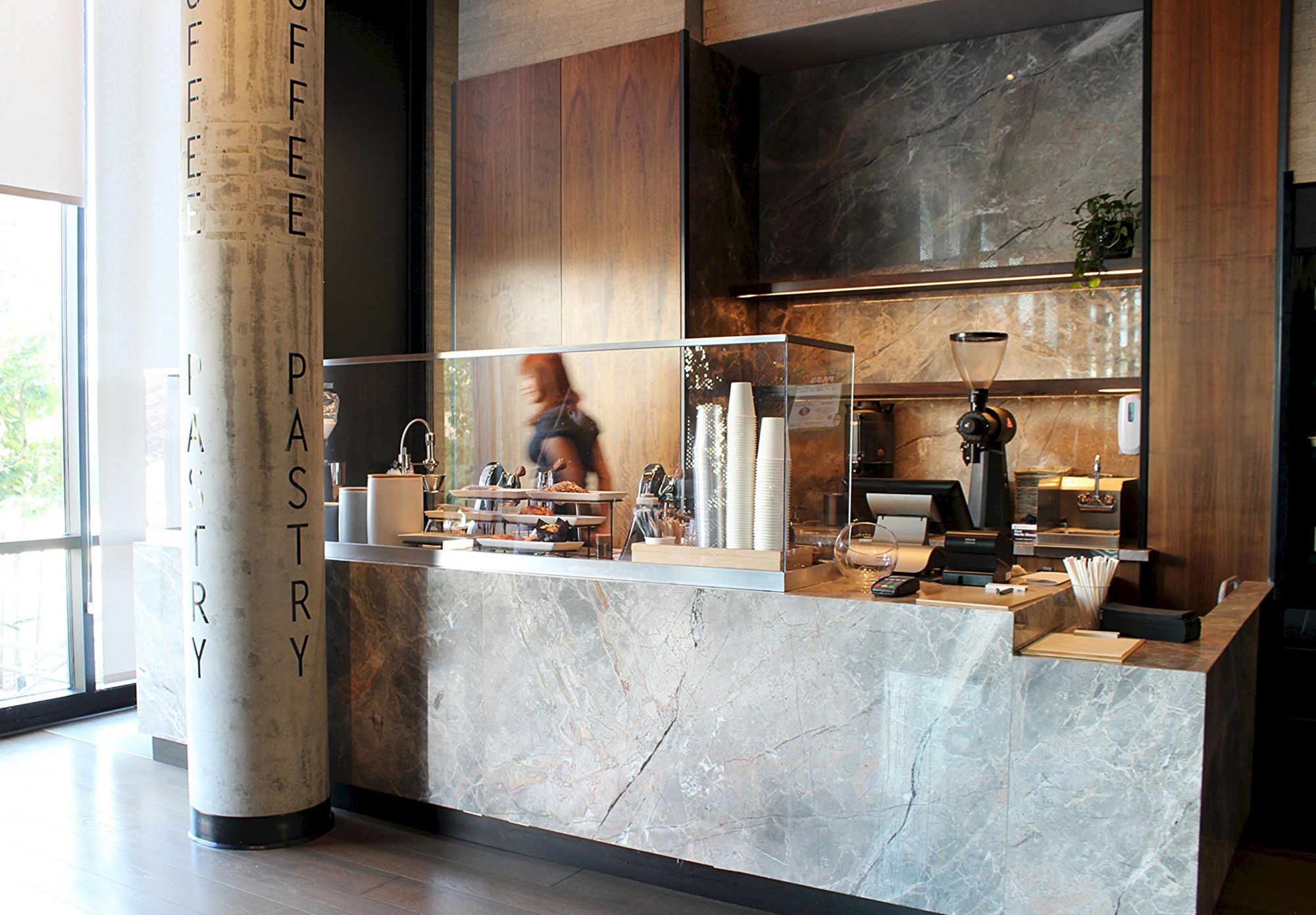 A modern cafe counter with pastries, coffee equipment, a cashier, and a person behind the counter, with wood and concrete design elements.