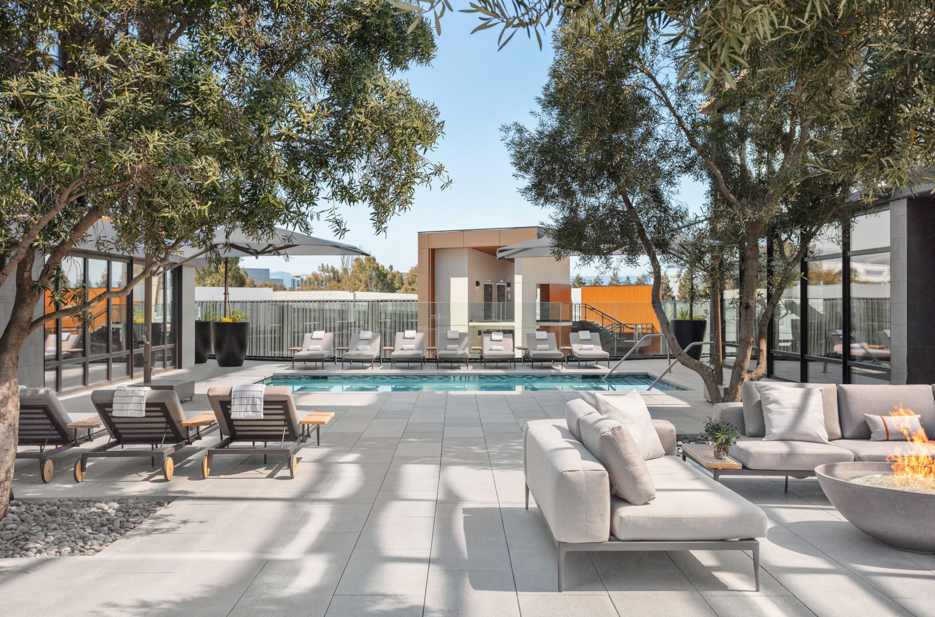 A modern outdoor space with a pool, sun loungers, sofas, trees, and a fire pit, surrounded by glass structures and greenery.