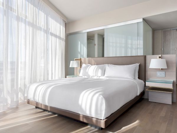 A bright hotel room with a large bed, bedside tables, lamps, and sunlight streaming through sheer curtains.