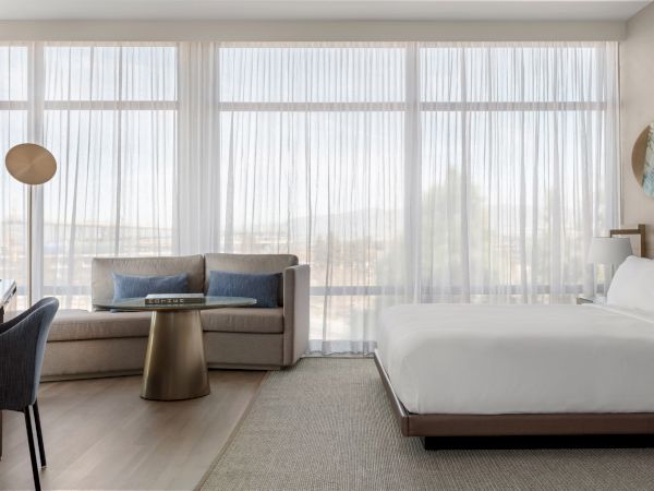 A modern hotel room with a cozy sofa, bed, desk, and lamp, featuring large windows with sheer curtains providing a bright, airy feel.