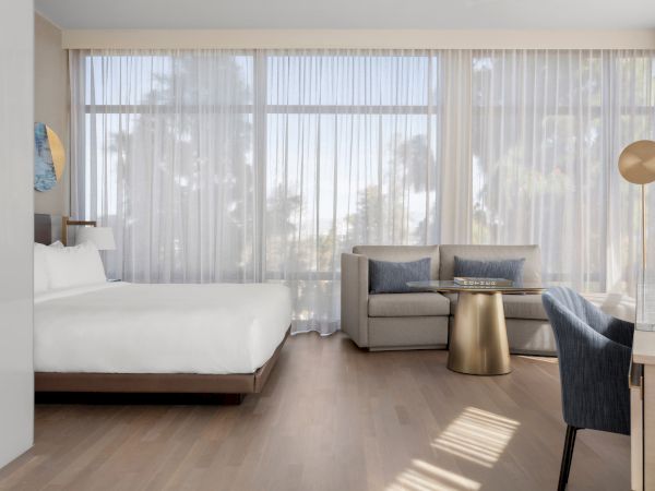 A modern hotel room with a bed, sofa, round table, and large windows with sheer curtains letting in natural light.
