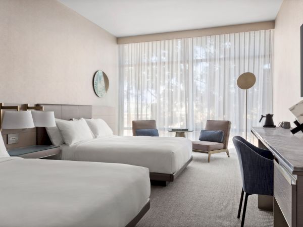 A modern hotel room with two beds, a desk, chair, and a sitting area by the window with sheer curtains and gentle lighting.
