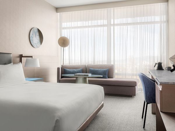 A modern hotel room with a bed, sofa, desk, and a large window with sheer curtains, creating a bright and cozy atmosphere.