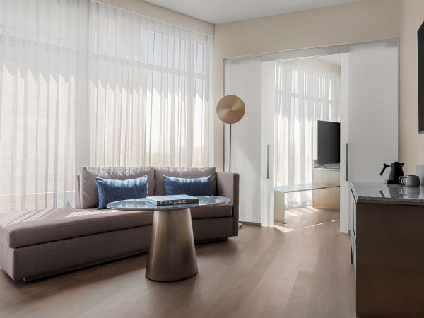 A modern living room with a sofa, blue pillows, round glass table, TV, sliding doors, sheer curtains, and a kettle on a counter.