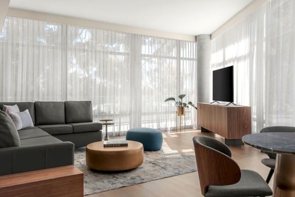 A modern living room with a dark sectional sofa, TV on a stand, round coffee table, and dining chairs, illuminated by natural light through sheer curtains.
