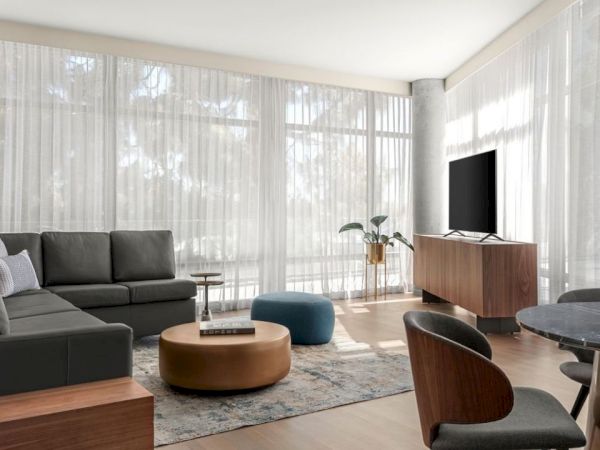 A modern living room with a dark sectional sofa, TV on a stand, round coffee table, and dining chairs, illuminated by natural light through sheer curtains.