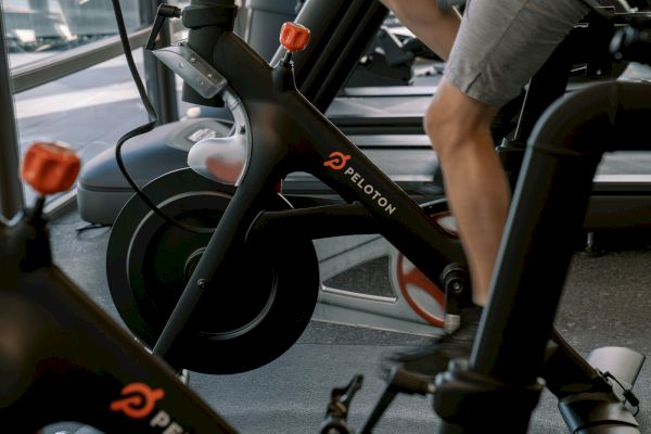 The image shows a person using a Peloton exercise bike in a gym setting.