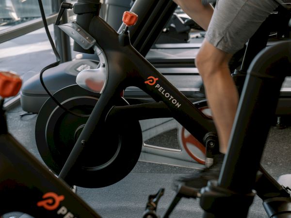 The image shows a person using a Peloton exercise bike in a gym setting.